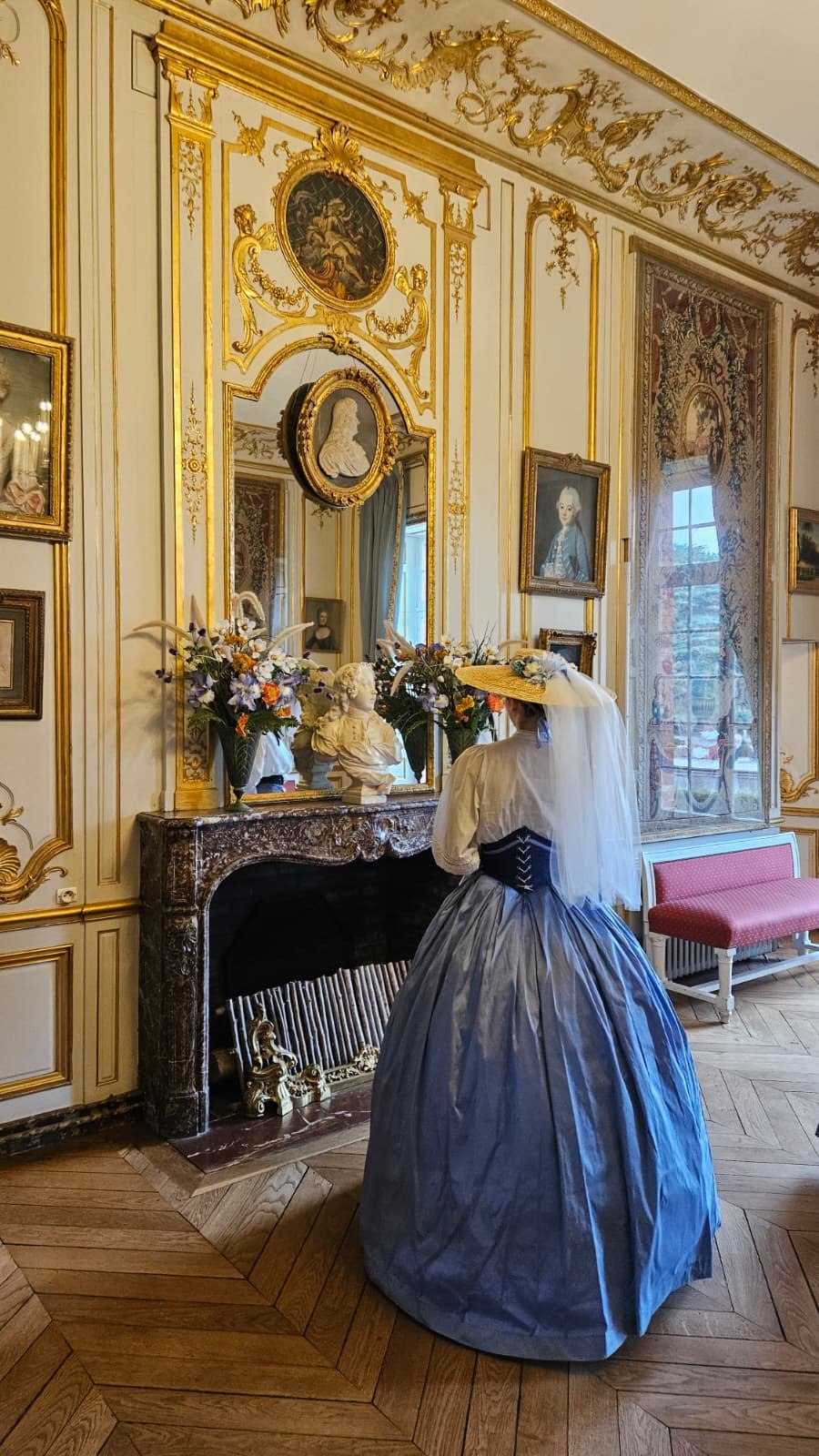 Participante en robe bleue à crinoline devant une cheminée et un miroir dans un salon du Château de Breteuil