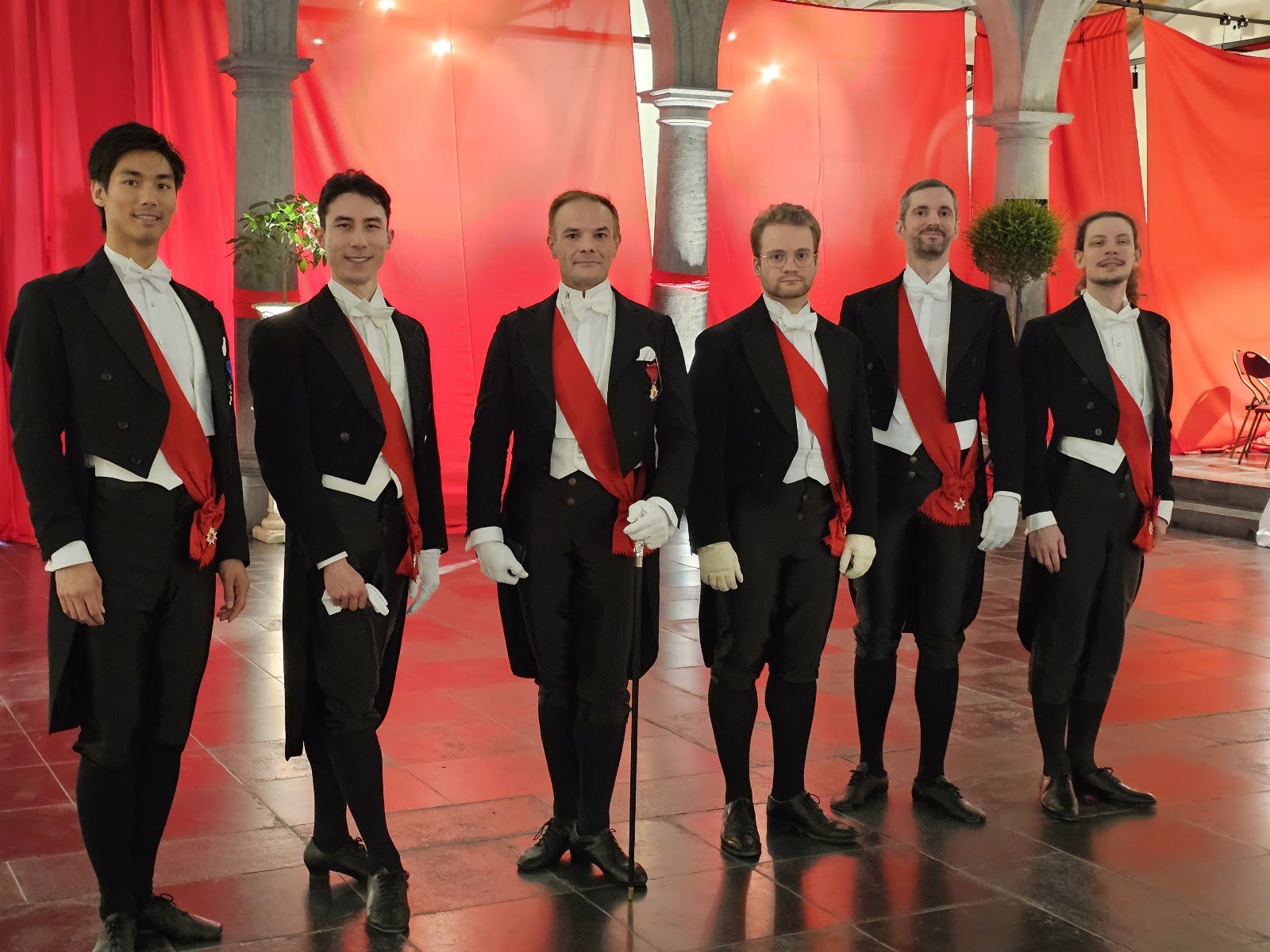 Groupe de danseurs en habits noirs et écharpes rouges devant les tentures du bal — Senso 1866, 26 avril 2025